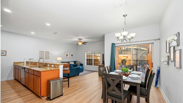 A modern kitchen with wooden cabinets and a stainless steel refrigerator, a dining area with a table and chairs, and a living room with a blue sofa and a ceiling fan. - Bright & Breezy Disney Escape | Pool | BBQ (Kissimmee)