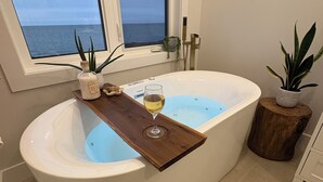 A modern bathroom with a freestanding bathtub, a wooden tray with a glass of wine, and a view of the sea through a window.