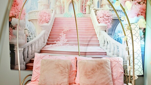 Un dormitorio con una cama con ropa de cama rosa y almohadas mullidas, un arco decorativo y un mural de una imponente escalera adornada con flores rosadas.