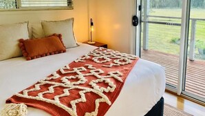 A bedroom with a bed, pillows, a nightstand, a lamp, and a view of a deck and greenery outside.
