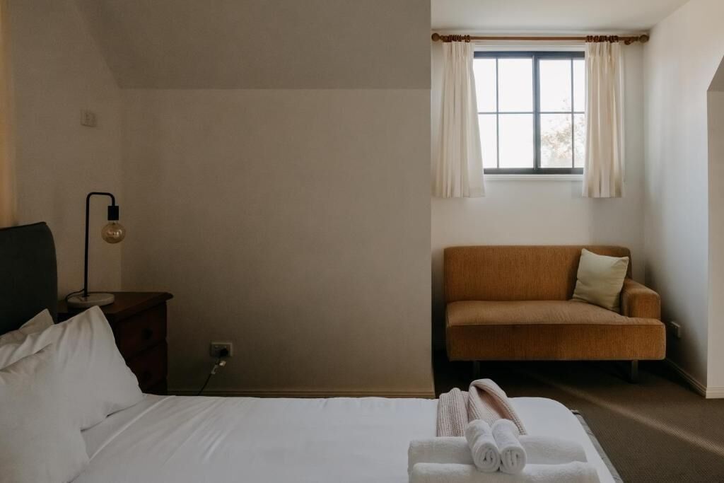 A hotel room with a bed, a nightstand, a lamp, a sofa, and a window with curtains.