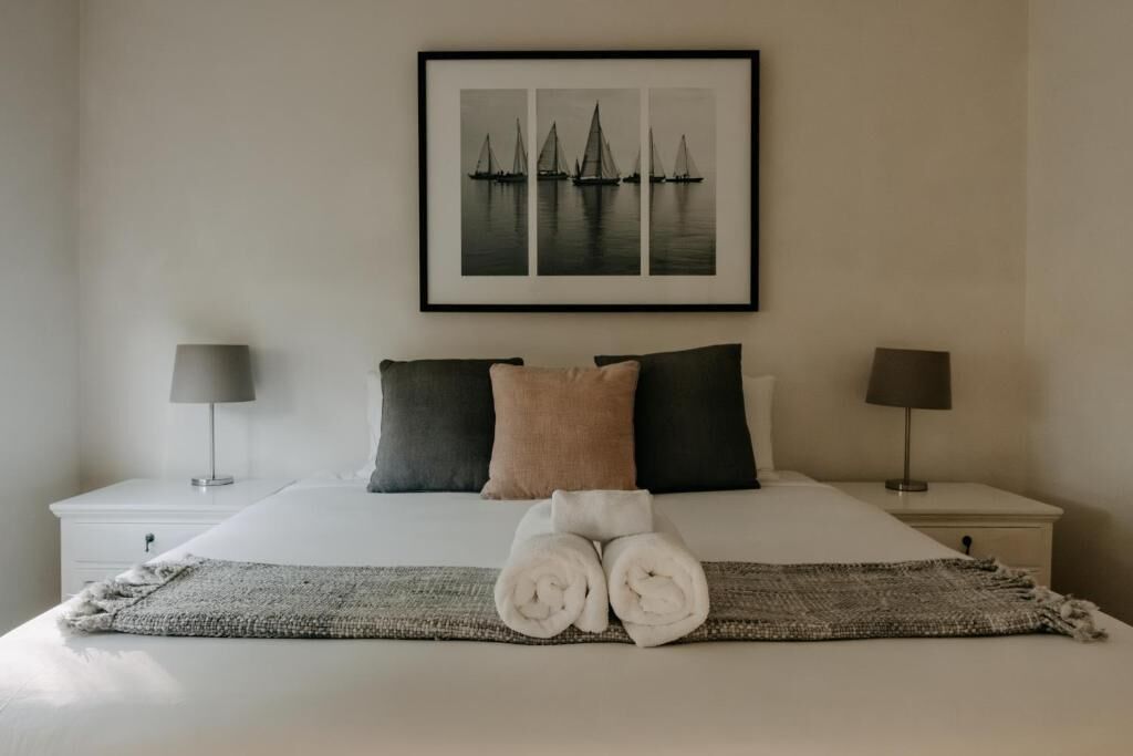 Un lit bien fait, avec des serviettes pliées, deux tables de chevet avec des lampes et, au mur, un tableau représentant des voiliers.