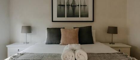 Un lit bien fait, avec des serviettes pliées, deux tables de chevet avec des lampes et, au mur, un tableau représentant des voiliers.