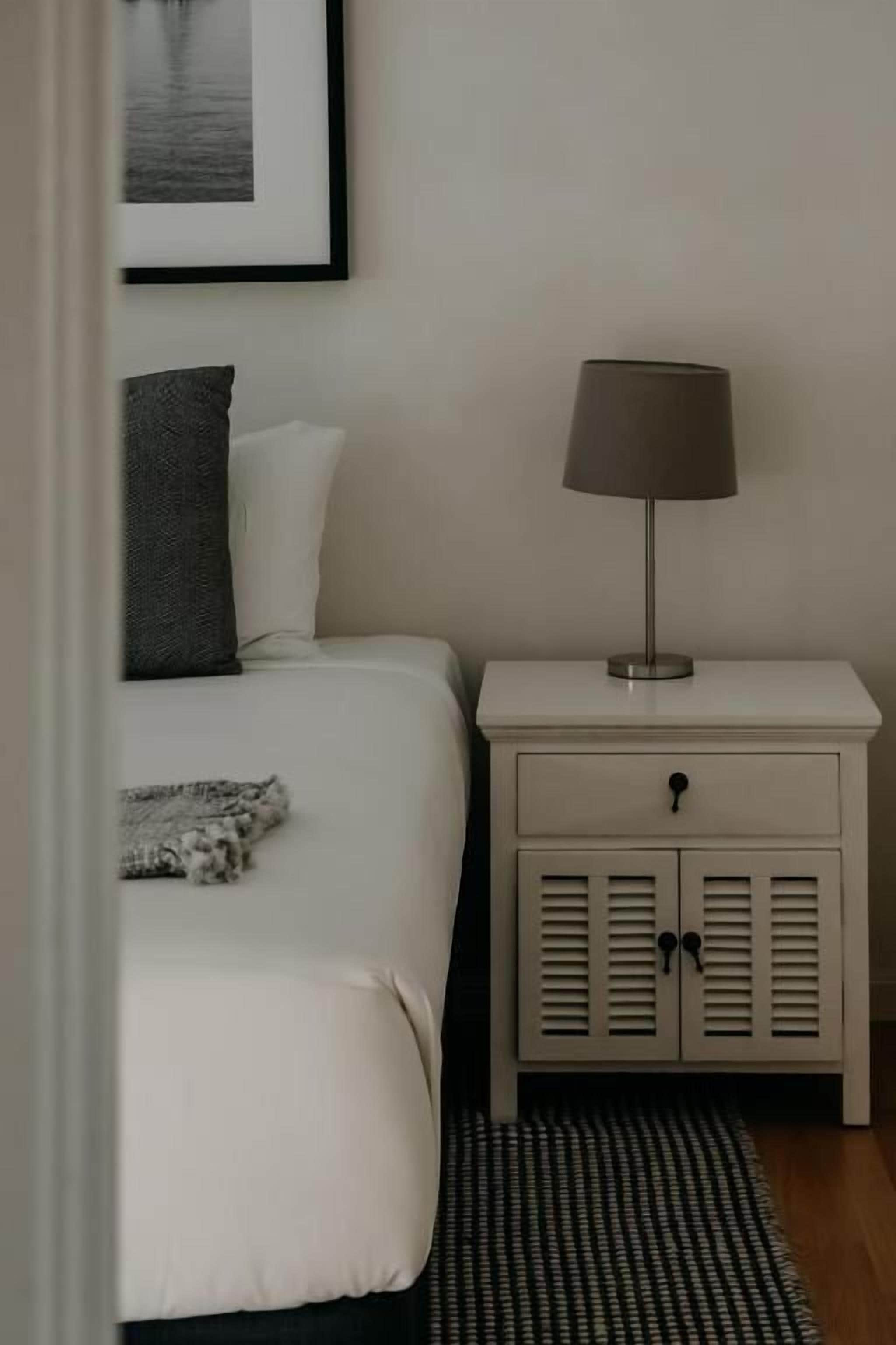 Un dormitorio con cama, una mesita de noche con lámpara, un cuadro enmarcado en la pared y una alfombra estampada.