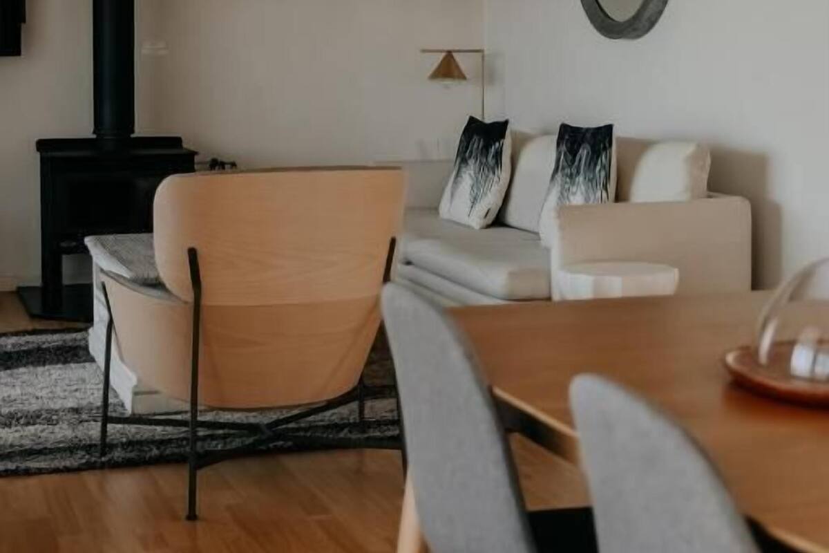 Ett modernt vardagsrum med en soffa, fåtöljer, ett matbord, en öppen spis och en spegel som hänger på väggen.
