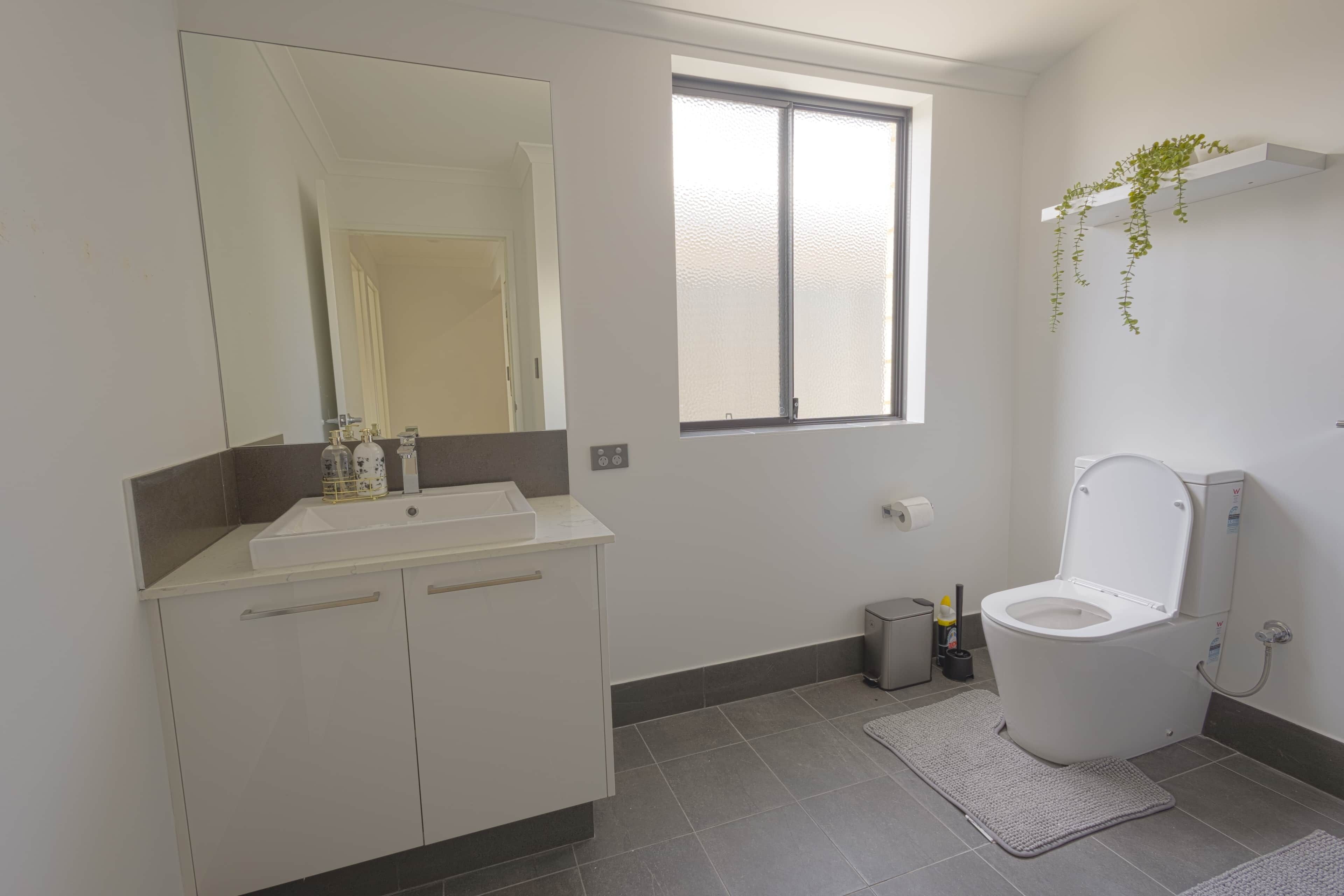 Un bany modern amb un lavabo, un mirall i un vàter de color blanc. Hi ha un finestral que aporta llum natural i un petit cendrer al costat del vàter.