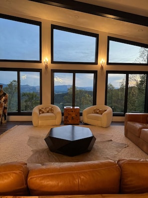 A modern living room with large windows, leather sofas, and a central coffee table. - Luxury Cabin w/ Epic Views + Hot Tub & Firepit (Bryson City)