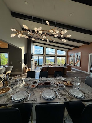 A modern dining area with a marble table set for a meal, large windows offering a view, and a unique ceiling light fixture. - Luxury Cabin w/ Epic Views + Hot Tub & Firepit (Bryson City)