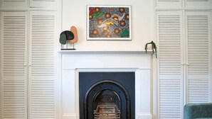 A living room with a fireplace, white shutters, a blue sofa, a wooden coffee table, and a framed artwork above the fireplace.