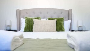 A neatly made bed with a grey upholstered headboard, two green pillows, and a white pillow. There are two bedside tables with lamps, and the room has a clean, minimalist design.