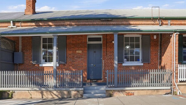 A two-story brick building with a grey metal roof, a front porch with a picket fence, and a blue door.
