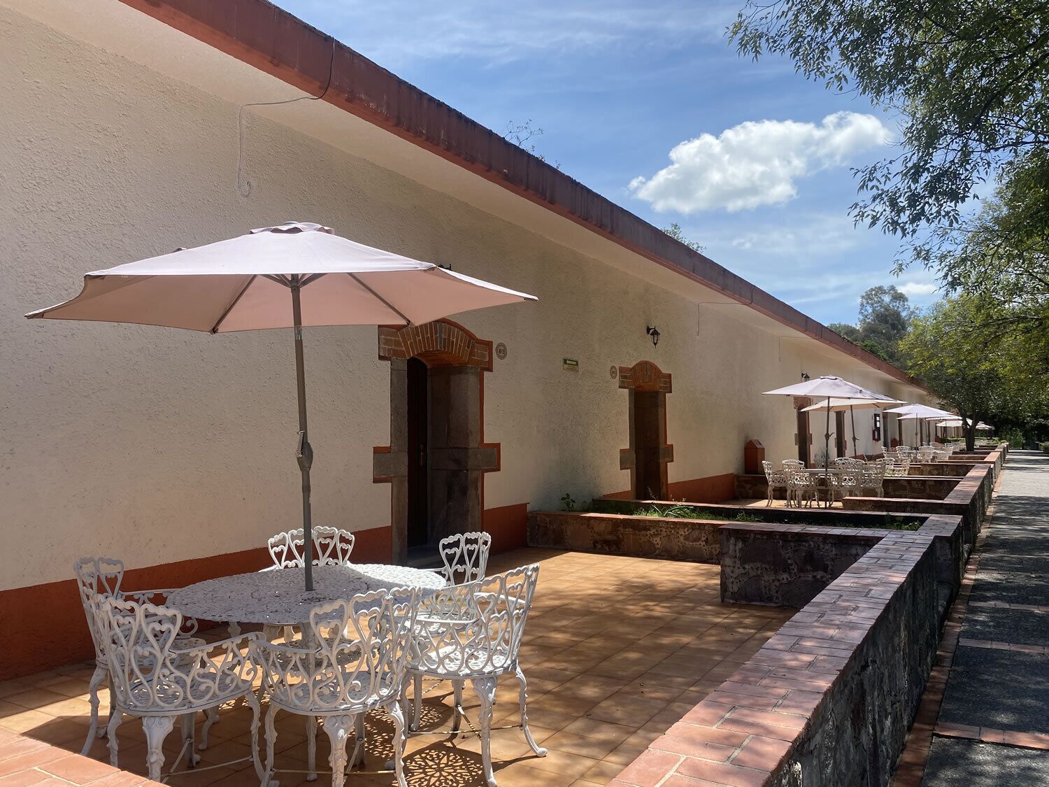 Une rangée de tables en plein air, avec des chaises blanches et des parasols, un chemin bordé de briques et un bâtiment à la façade beige et aux ornements rouges.