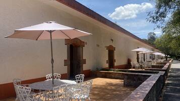 Une rangée de tables en plein air, avec des chaises blanches et des parasols, un chemin bordé de briques et un bâtiment à la façade beige et aux ornements rouges.