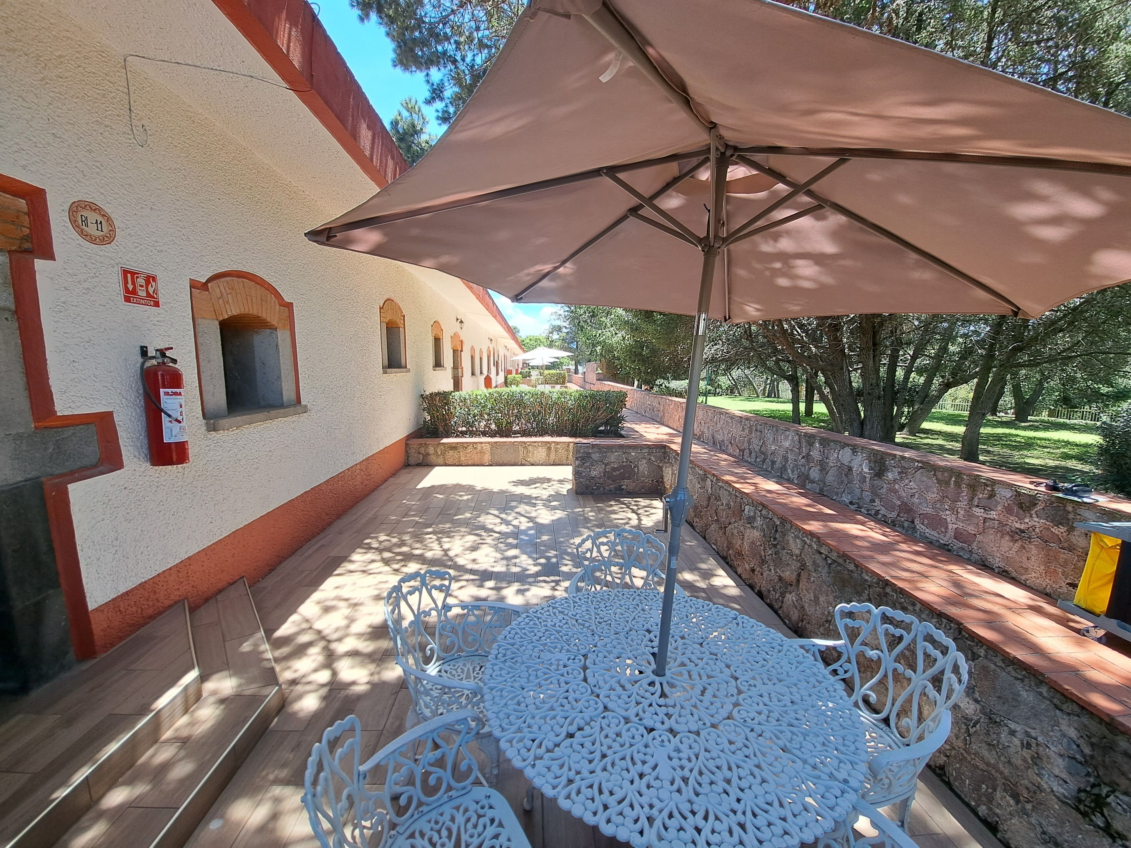 An outdoor patio with white metal tables and chairs, a large umbrella, and a fire extinguisher mounted on the wall.
