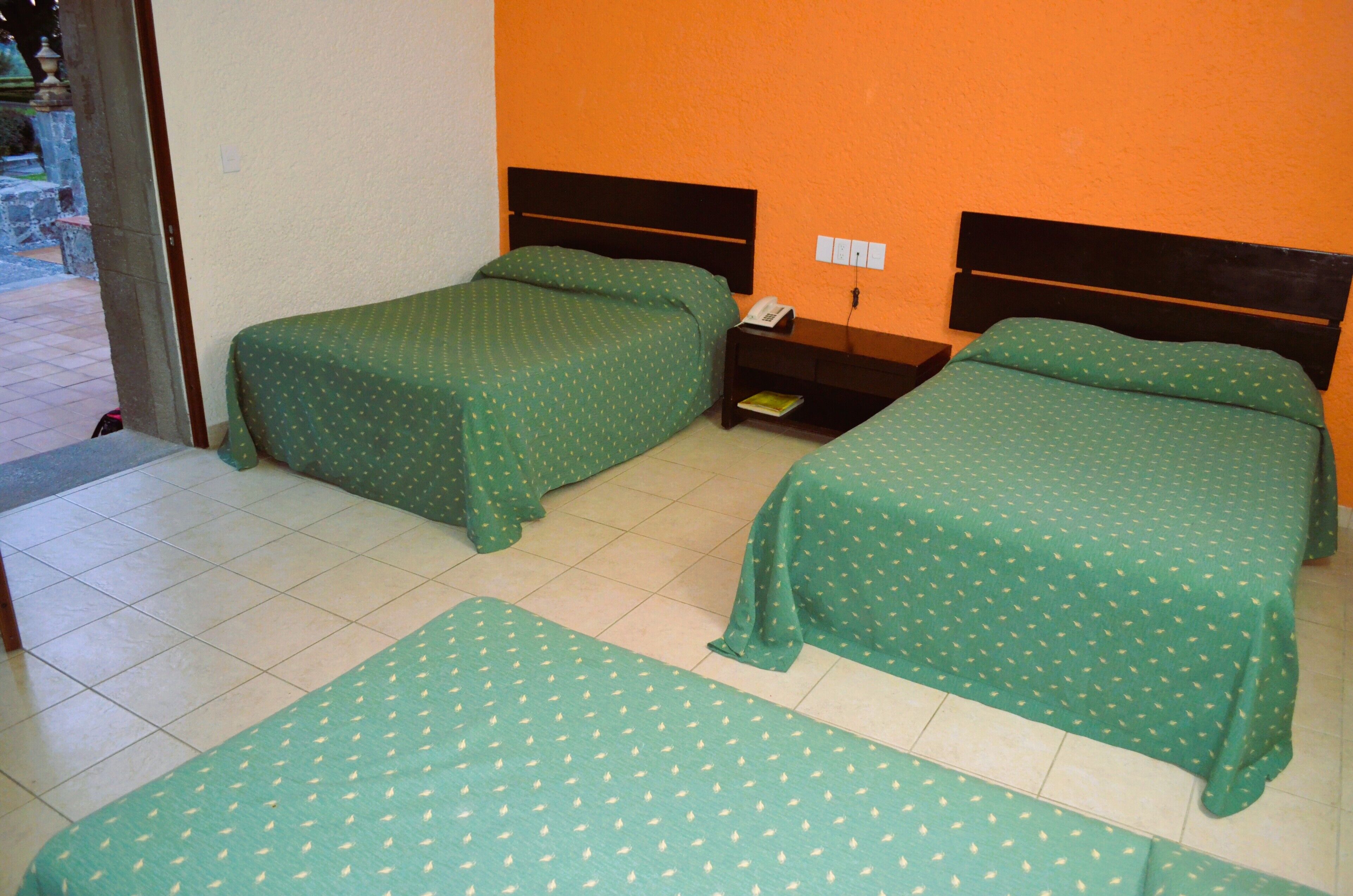 Una habitación con dos camas, cada una con ropa de cama verde, una mesita de noche con teléfono y suelo de baldosas.