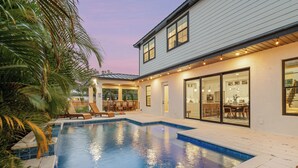 Una casa moderna con piscina, un patio con sillones de descanso y una cocina amplia visible a través de puertas de cristal.
