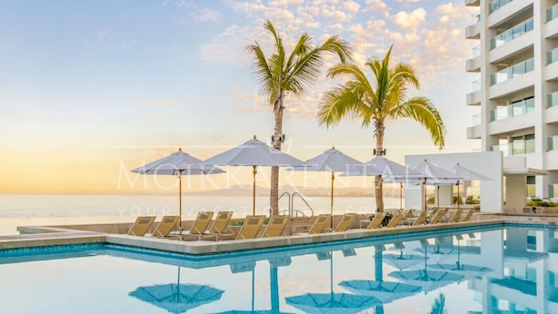 Ein Poolbereich mit Liegestühlen und Sonnenschirmen, Palmen und Blick auf das Meer bei Sonnenuntergang.