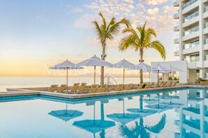 A poolside area with lounge chairs and umbrellas, palm trees, and a view of the ocean at sunset. - Oceanfront Condo | Sunset Views | 500Mbps WiFi (Puerto Vallarta)