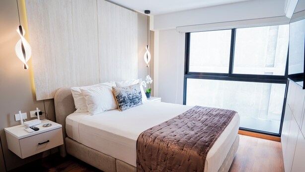 A neatly made bed with a brown quilt, a bedside table with a lamp, and a window with blinds. -  Miraflores Prime Apartment-3 blocks to Kennedy Park (Miraflores)