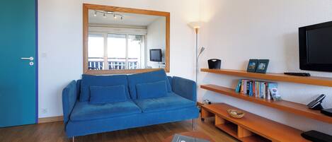 A modern living room with a blue sofa, wooden shelves, a TV, and a laptop on a round table.