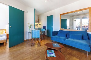 A modern living room with a blue sofa, wooden flooring, a dining area with chairs, and a kitchen in the background. - Appartement Lilas - Welkeys (Soorts-Hossegor)