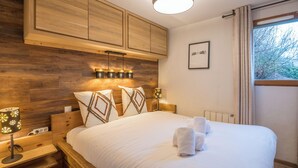A bedroom with a wooden headboard, a bed with white linens, a bedside table with a lamp, and a window with curtains. - Appartement Rocca - Welkeys (Saint-Gervais-les-Bains)