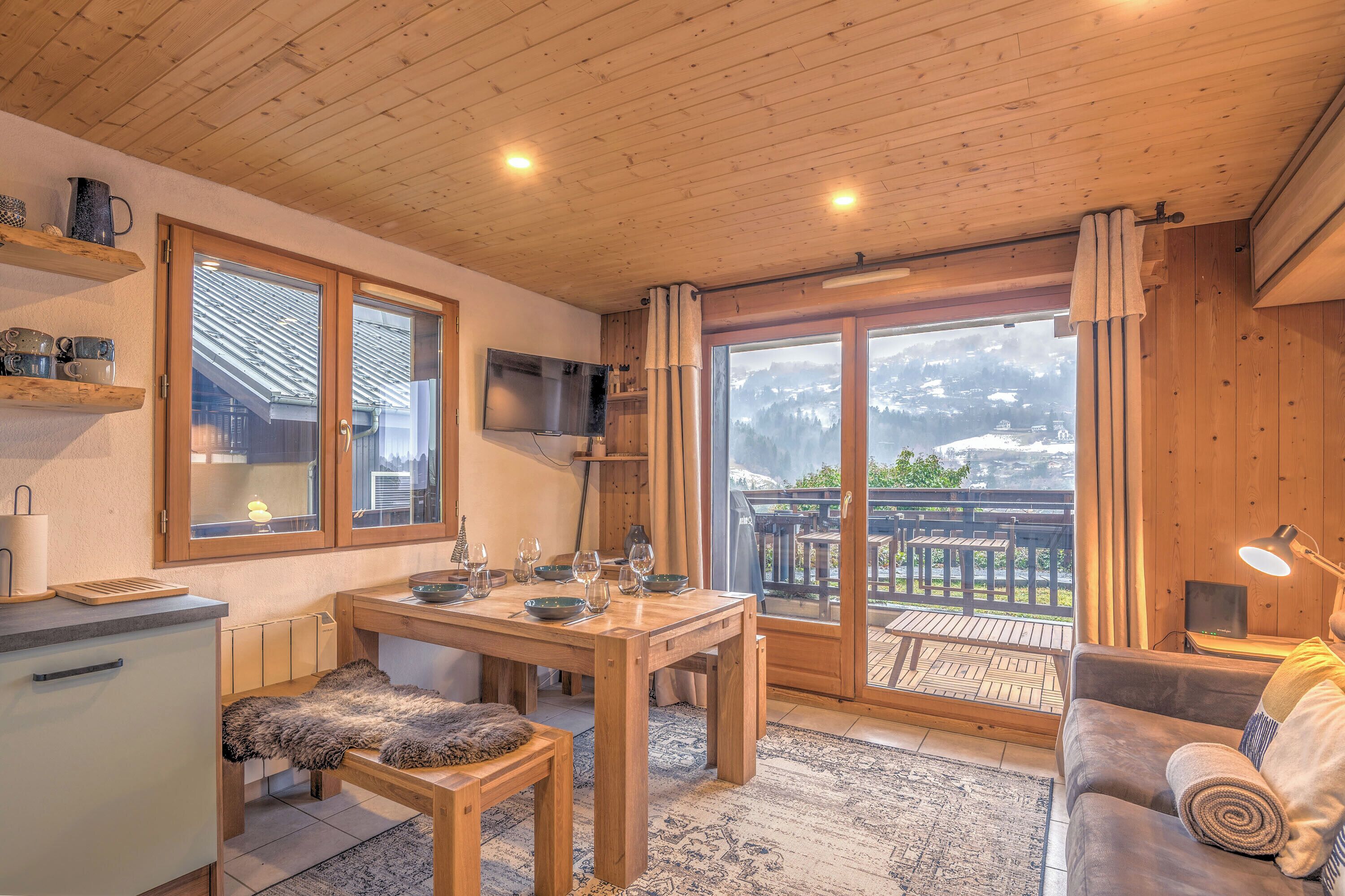 Een gezellig houten interieur met een eettafel gedekt voor vier personen, een bank met een opgerolde deken, en een groot raam met uitzicht op een besneeuwde berg.