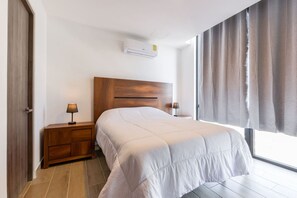 Habitación de hotel con cabecera de madera, cama con edredón blanco, dos mesitas de noche con lámparas, ventana con cortinas y un equipo de aire acondicionado en la pared.