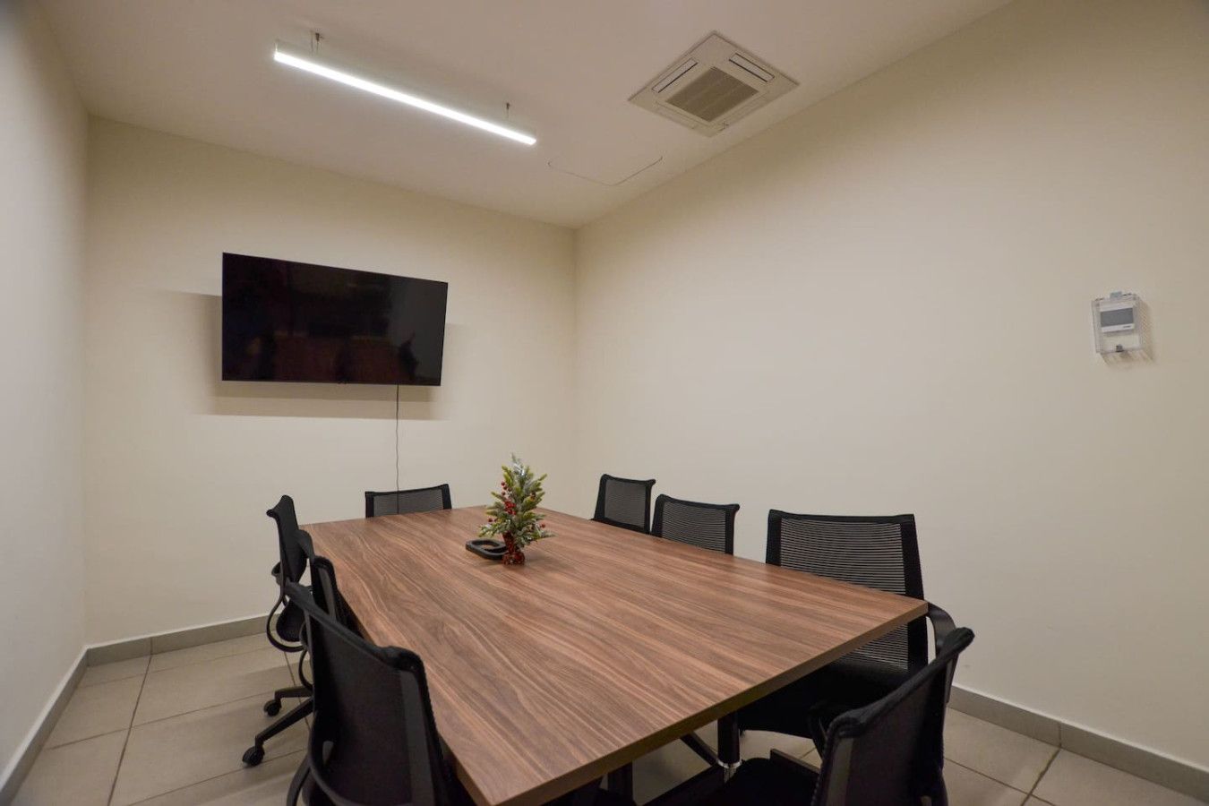 A conference room with a long table, black chairs, a small Christmas tree, a flat-screen TV, and a ceiling vent.