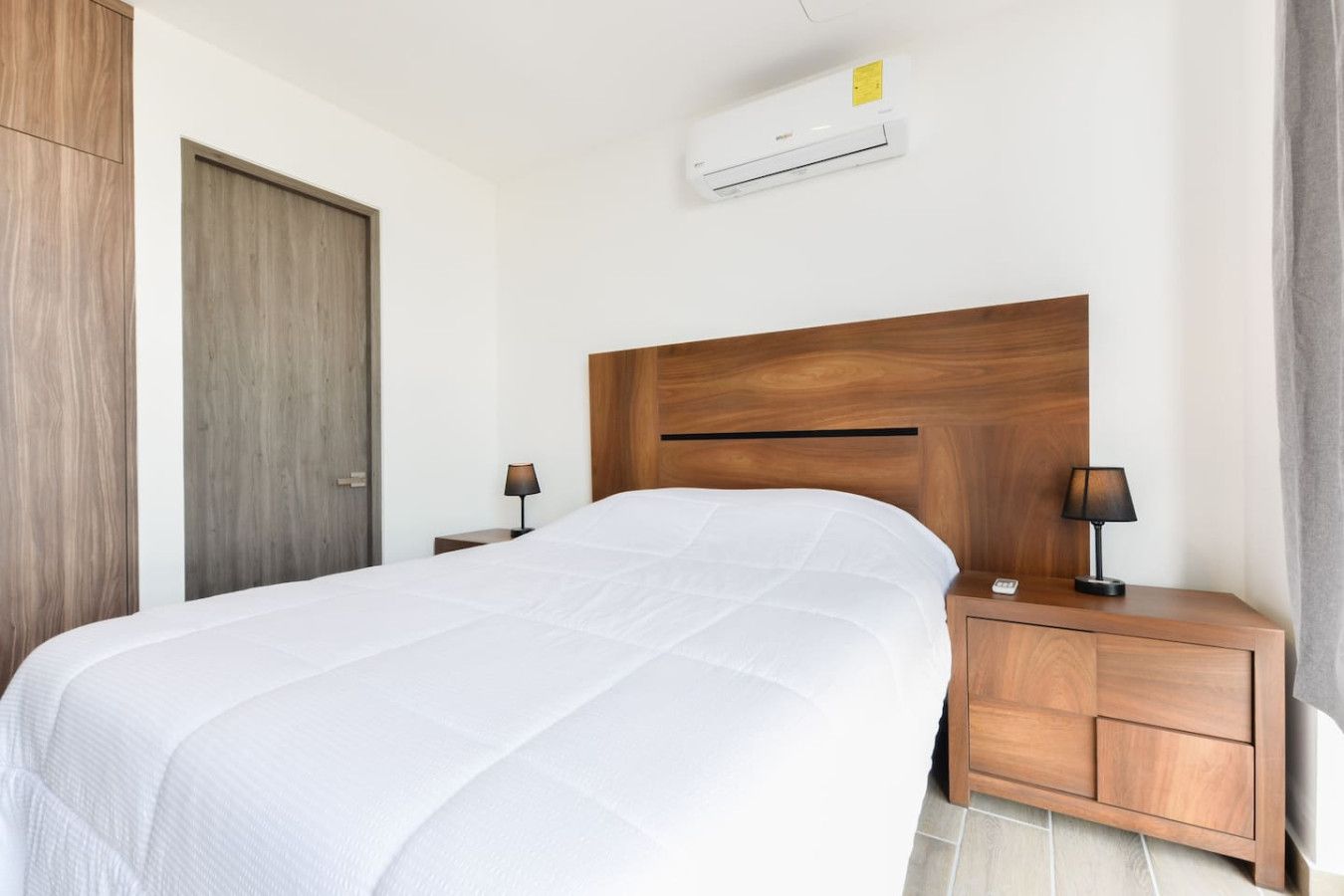 Een net opgemaakt bed met een houten hoofdbord, een nachtkastje met een lamp en een airconditioner aan de muur.