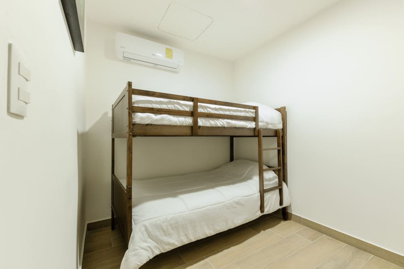 Un lit superposé dans une pièce avec un mur blanc, une bouche d’aération au plafond et un interrupteur.