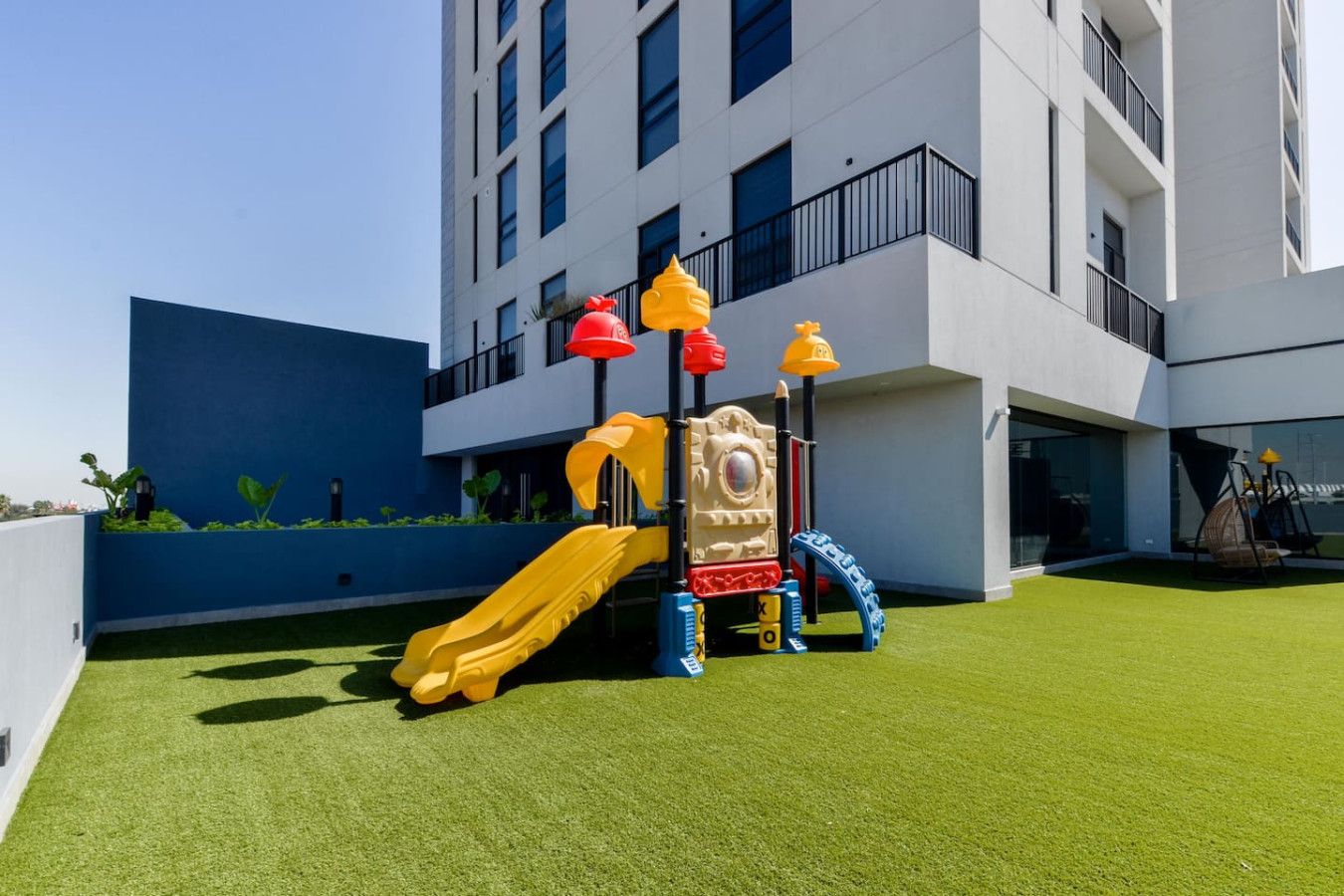 Un terrain de jeux équipé d’un toboggan et de structures d’escalade, situé devant un bâtiment moderne doté de balcons.