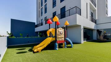 Un terrain de jeux équipé d’un toboggan et de structures d’escalade, situé devant un bâtiment moderne doté de balcons.