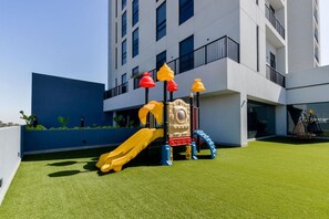 Een speeltuin met een glijbaan en klimconstructies, gelegen voor een modern gebouw met balkons.