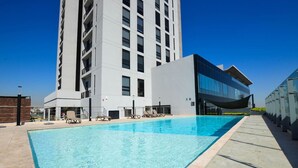 Een modern hotel met een zwembad, ligbedden en een hoog gebouw op de achtergrond.