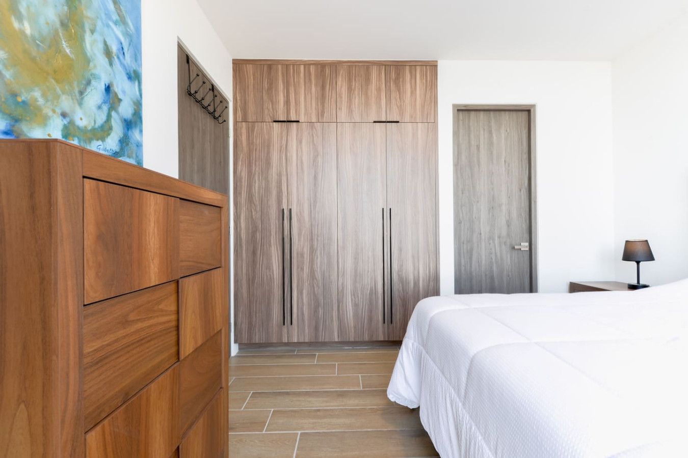 A bedroom with a wooden wardrobe, a bed with white bedding, a nightstand with a lamp, and a door with a wooden panel.