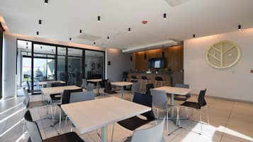 A modern dining area with tables and chairs, a kitchen area, and large windows allowing natural light.