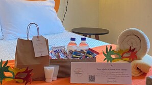 Un lit bien fait, avec un oreiller blanc et une tête de lit orange. Une petite table avec une lampe se trouve dans un coin. Un sac cadeau, des bouteilles d’eau et une carte de bienvenue sont posés sur le lit.
