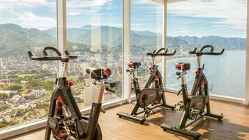 Três bicicletas ergométricas em um ambiente com amplas janelas com vista para uma cidade e montanhas.