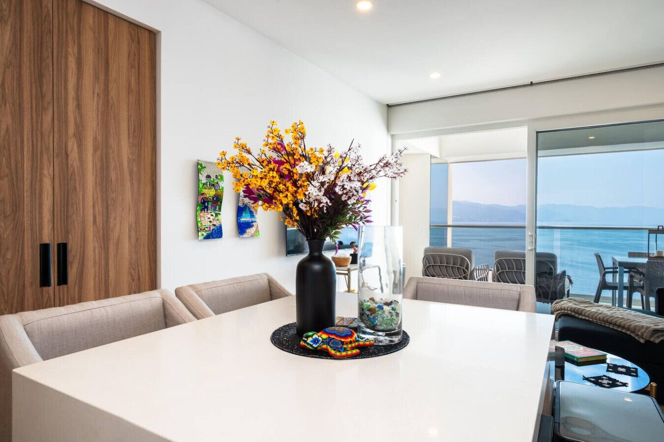 Uma sala de jantar moderna com uma mesa branca, flores coloridas e vista para o mar.