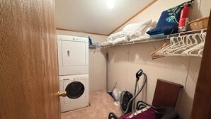 A laundry room with a front-loading washer and dryer, a vacuum cleaner, and a storage area with shelves and hanging clothes. - Charming 3 Bed Room Monthly Rental in the Heart of Ellicottville (Ellicottville)