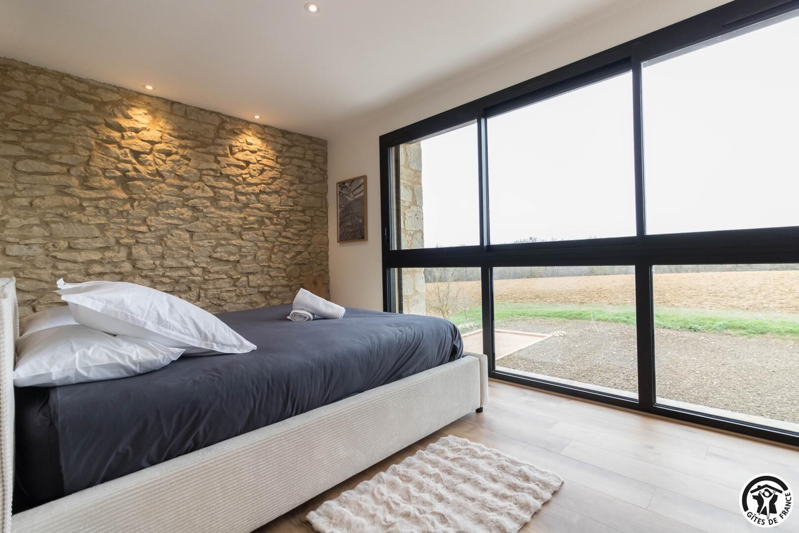 Ein Schlafzimmer mit Steinwand, einem großen Bett mit weißer und grauer Bettwäsche sowie Blick auf ein Feld durch große Fenster.