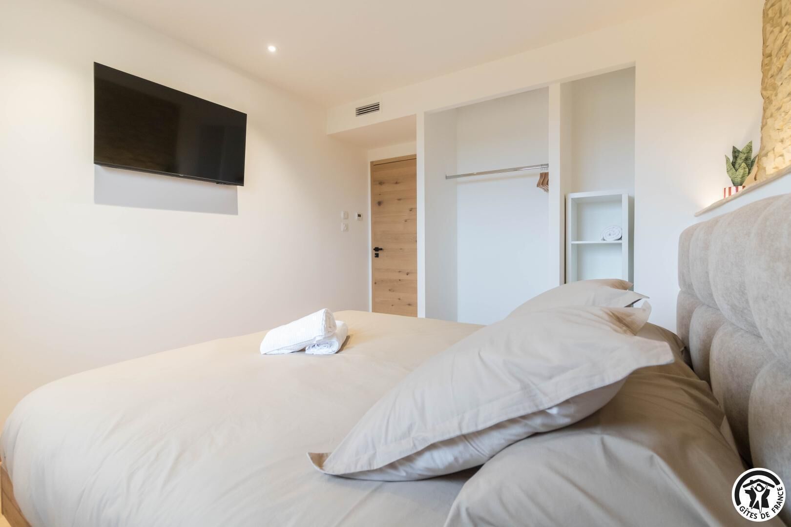 A modern bedroom with a large bed, a flat-screen TV mounted on the wall, and a wooden door leading to a walk-in closet.