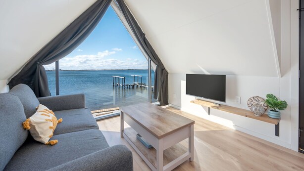 A modern living room with a large window overlooking a body of water, a sofa with cushions, a wooden coffee table, a flat-screen TV, and a shelf with decorative items. - Waterfront A-Frame Panoramic Sunset Views (Fort Walton Beach)