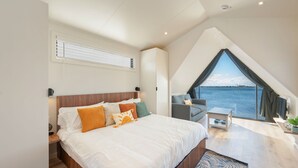 A modern bedroom with a large bed, a small seating area, and a view of the water. - Waterfront A-Frame Panoramic Sunset Views (Fort Walton Beach)