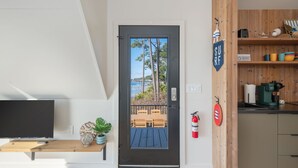 A modern living room with a TV, a wooden bench, a plant, and a door leading to an outdoor seating area with a view of trees and water. - Waterfront A-Frame Panoramic Sunset Views (Fort Walton Beach)