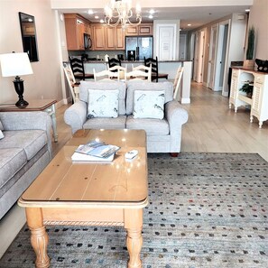 A living room with a sofa, coffee table, and a rug. There is a kitchen in the background with wooden cabinets and a chandelier. - 204 - Beachfront Paradise On The Second Floor! (Panama City Beach)