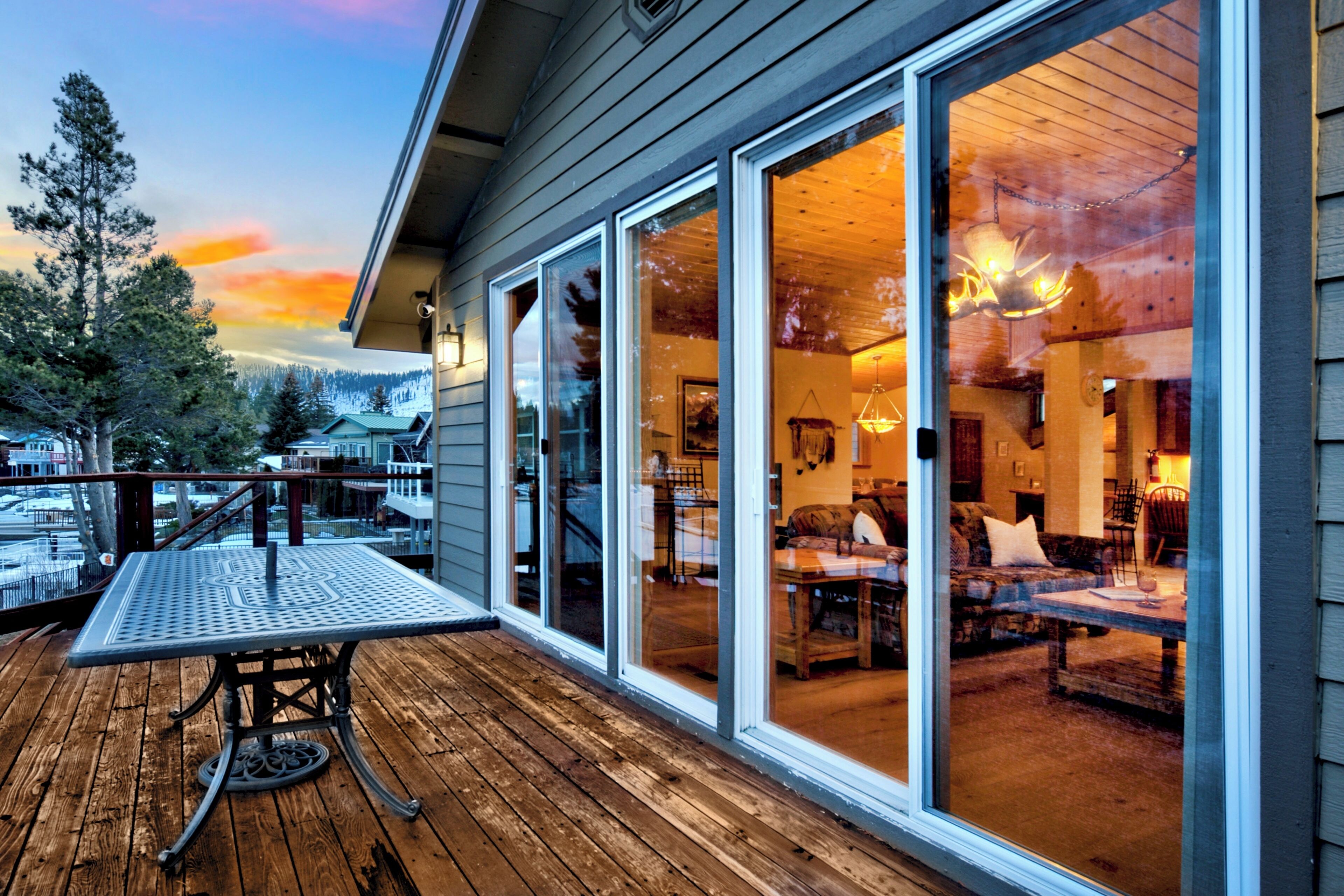 Una terraza de madera con una mesa y sillas de metal, amplias puertas correderas de vidrio que dan a un interior con chimenea y zona de estar.