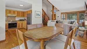 A spacious kitchen with wooden cabinets, a dining table with chairs, a refrigerator, and a stove. There is a staircase leading to a living area with a sofa and a TV. - The Aspen Hideaway | Elkhorn Pool, Hot Tub + Pickleball (Sun Valley)