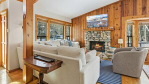A cozy living room with a stone fireplace, wooden walls, and a large window overlooking a snowy landscape. - The Aspen Hideaway | Elkhorn Pool, Hot Tub + Pickleball (Sun Valley)